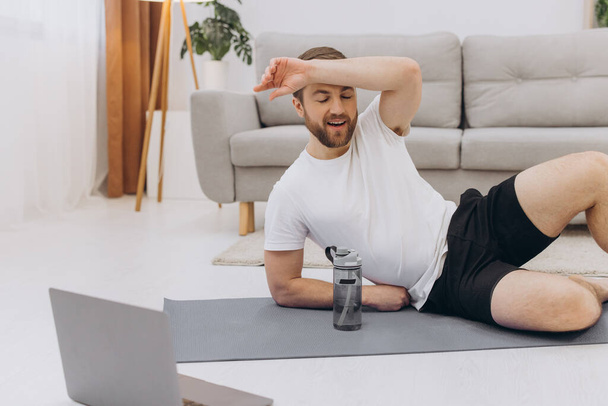 Handsome adult man using laptop and drinking water while having break during workout at home - Photo, Image
