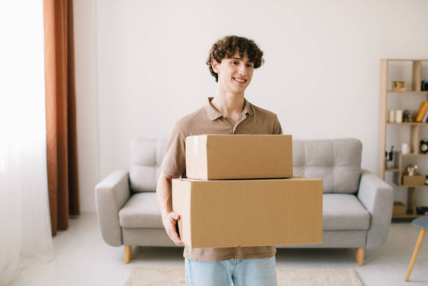 Shopping And Delivery. Curly Young Man Customer Holding Cardboard Box Posing Smiling To Camera Standing At Home. Excited Male Buyer Hugging Delivered Parcel. - Photo, Image