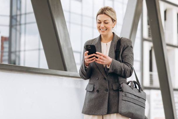 Porträt einer schönen reifen Geschäftsfrau im Anzug und grauer Jacke, die vor dem modernen urbanen Hintergrund lächelt und telefoniert - Foto, Bild
