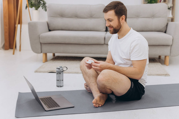 Vonzó szakállas férfi kezében vezeték nélküli fejhallgató közelében laptop és kezdő reggeli testmozgás vagy jóga. - Fotó, kép