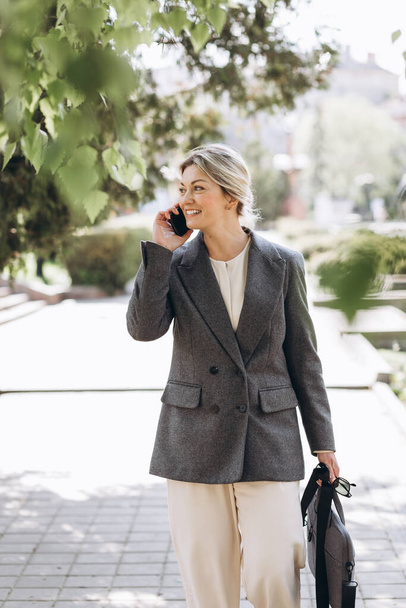Зрелая блондинка, улыбающаяся деловая женщина ходит по улицам города и говорит по телефону - Фото, изображение