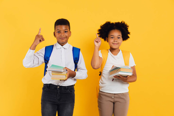 Happy african american schoolgirl and schoolboy holding books and showing something together on yellow background, copy space. - Photo, Image