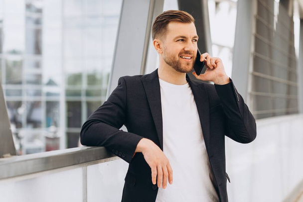 Egy jóképű, boldog férfi öltönyös üzletember portréja, aki telefonon beszél a városi épületek és irodák hátteréről.. - Fotó, kép