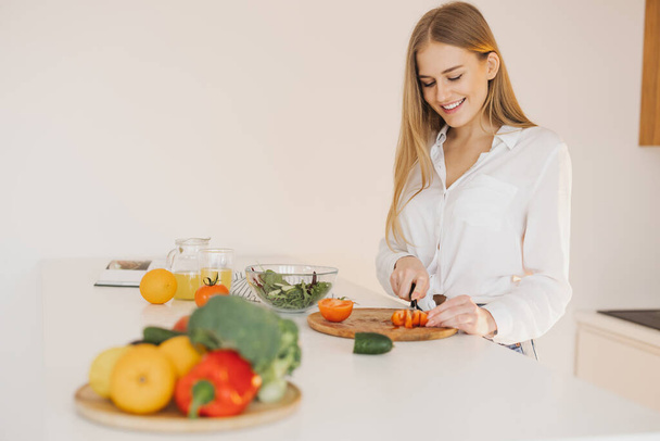 Una mujer rubia linda y feliz está preparando una ensalada en la cocina - Foto, imagen