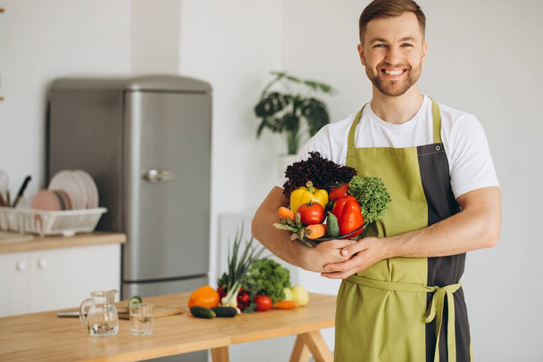 Πορτρέτο ενός ευτυχισμένου άνδρα κρατώντας ένα πιάτο φρέσκα λαχανικά στο φόντο της κουζίνας στο σπίτι - Φωτογραφία, εικόνα