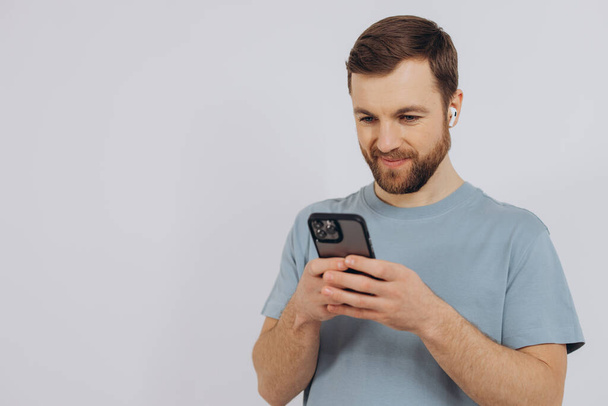 Портрет современного человека тысячелетия с бородой, держащего смартфон и слушающего что-то в беспроводных наушниках на белом фоне с копировальным пространством - Фото, изображение