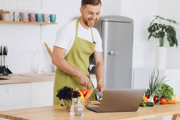 Portrait of happy man at home, man cooking vegetable salad looking at camera and smiling, slicing vegetables, using laptop for online cooking training - 写真・画像