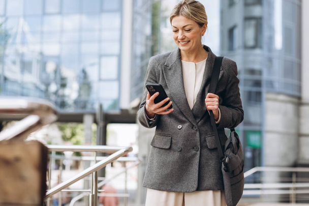 Retrato de una hermosa mujer de negocios madura en traje y chaqueta gris sonriendo y hablando por teléfono en el fondo urbano moderno - Foto, imagen