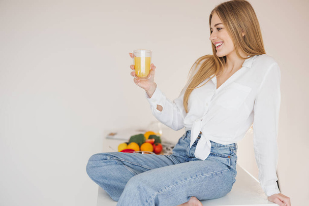 Happy and smiling blonde woman sitting on top of kitchen table and drinking orange juice among cooking ingredients. - Foto, Bild