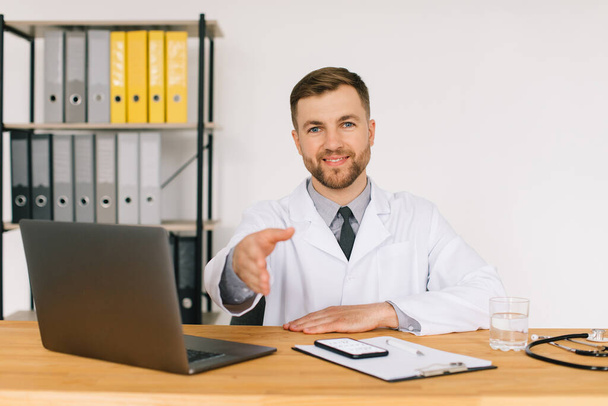 Feliz médico masculino viendo un webinar médico en línea o seminario de capacitación mientras está sentado con una computadora portátil en el lugar de trabajo. - Foto, Imagen