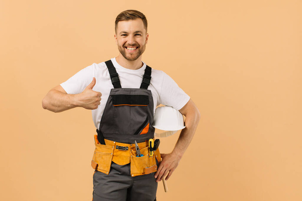 Positive männliche Reparaturarbeiter zu Hause hält einen Helm und zeigt perfekt auf einem beigen Hintergrund - Foto, Bild