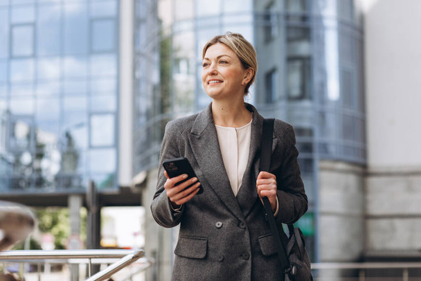 Ritratto di una bella donna d'affari matura in abito e giacca grigia che sorride e parla al telefono sullo sfondo urbano moderno - Foto, immagini