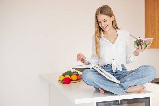 Szczęśliwa i uśmiechnięta blondynka siedząca na stole w kuchni i czytająca książkę kucharską oraz degustująca sałatkę domowej roboty wśród składników kuchennych. - Zdjęcie, obraz