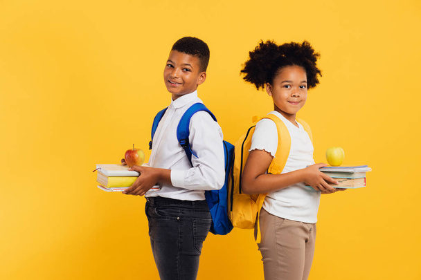 Fröhliches afrikanisch-amerikanisches Schulmädchen und Schüler mit gemischter Rasse, die nebeneinander Bücher auf gelbem Hintergrund halten. Zurück zum Schulkonzept. - Foto, Bild