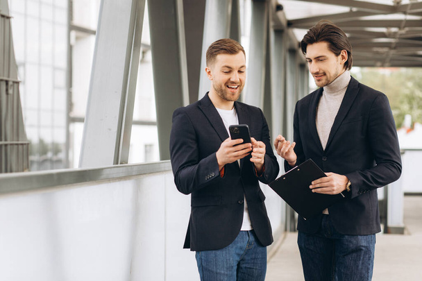 Dos hombres de negocios modernos están discutiendo algo y mostrándose mutuamente en un teléfono inteligente en el contexto de oficinas y edificios urbanos - Foto, imagen