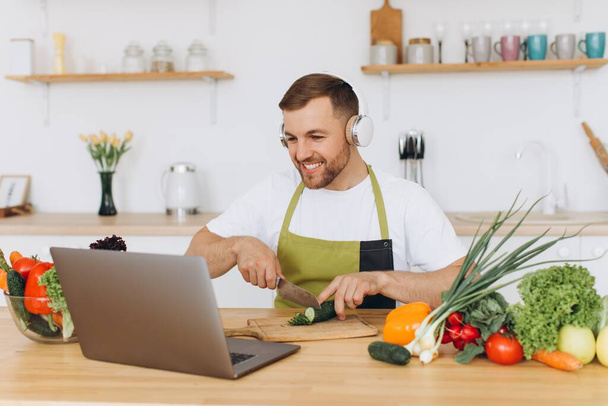 Szczęśliwy człowiek w słuchawkach przygotowuje sałatkę w kuchni ze świeżych warzyw i patrząc na laptopa. - Zdjęcie, obraz