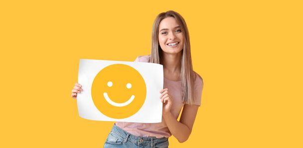 Kaunis nuori nainen pitää paperia hymyilevällä hymiöllä keltaisella taustalla - Valokuva, kuva