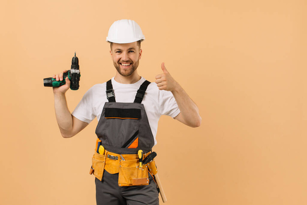 Позитивный мужчина-ремонтник дома держит отвёртку и хорошо показывает себя на бежевом фоне - Фото, изображение