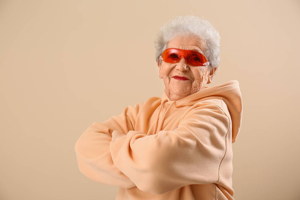 Donna anziana in occhiali da sole e felpa con cappuccio su sfondo beige - Foto, immagini