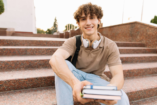 Estudante do sexo masculino atraente senta-se nos passos da universidade e lê algo, preparando-se para exames - Foto, Imagem