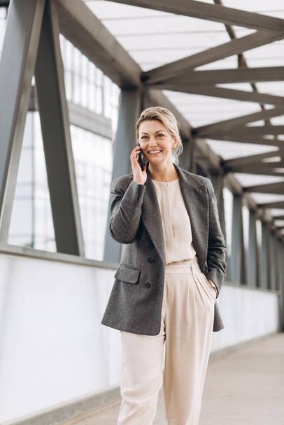 Portret pięknej dojrzałej kobiety biznesu w garniturze i szarej kurtce uśmiechającej się i rozmawiającej przez telefon na nowoczesnym tle budynków miejskich i biurowych - Zdjęcie, obraz