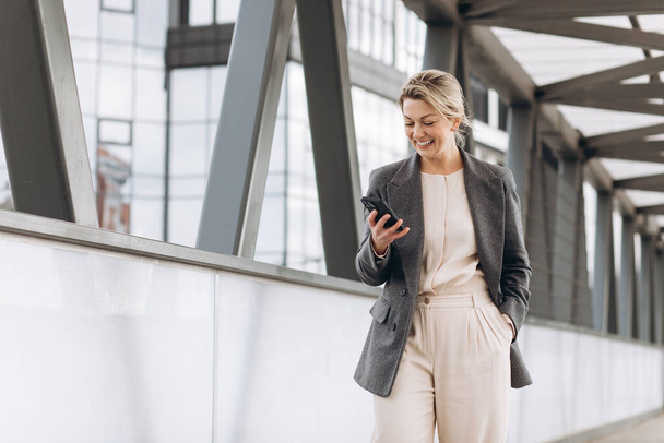 Porträt einer schönen reifen Geschäftsfrau in Anzug und grauer Jacke, die vor dem Hintergrund moderner Stadt- und Bürogebäude lächelt und telefoniert - Foto, Bild