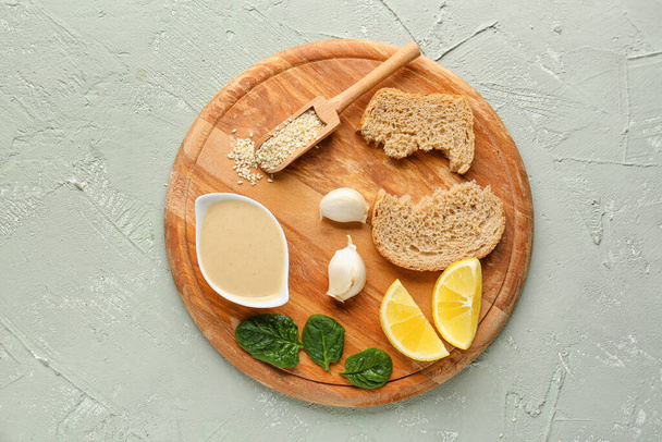 Wooden board with bowl of tasty tahini, sesame seeds, bread, garlic and lemon slices on grunge background - Фото, изображение