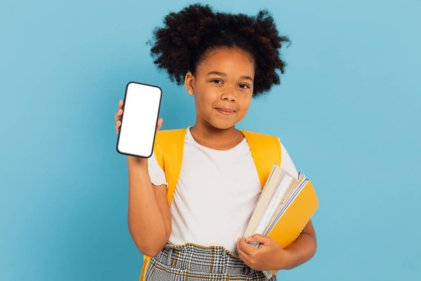 Petite fille afro-américaine excitée tenant une grande cellule à la main présentant un écran vide de téléphone sur fond de studio bleu. Maquette d'affichage pour smartphone - Photo, image
