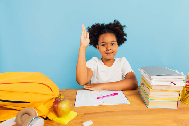 Fröhliches afroamerikanisches Schulmädchen sitzt im Unterricht am Schreibtisch und streckt die Hand vor blauem Hintergrund bergauf. Zurück zum Schulkonzept. - Foto, Bild