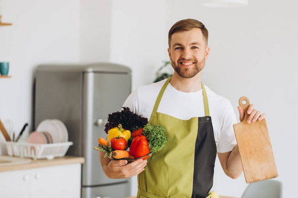 Portrait of happy man holding kitchen board and plate of fresh vegetables at home. - Photo, Image