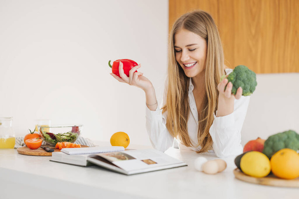 Portrait of a smiling cute blonde girl holding bell pepper and broccoli and reading recipe book in kitchen on table among cooking ingredients. - Foto, Imagem