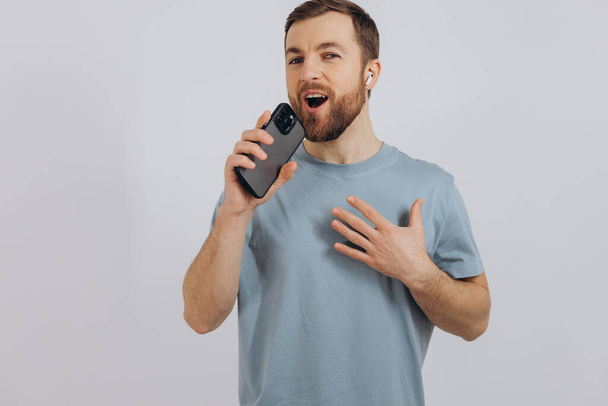 Happy bearded man dancing with earphones, listening music in headphones, holding mobile phone and smiling pleased, standing over white background - Foto, afbeelding