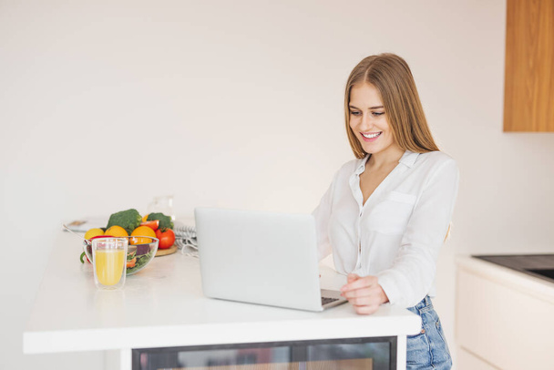 Glückliche und lächelnde blonde Frau auf der Suche nach Rezept am Laptop im Internet in der Küche - Foto, Bild