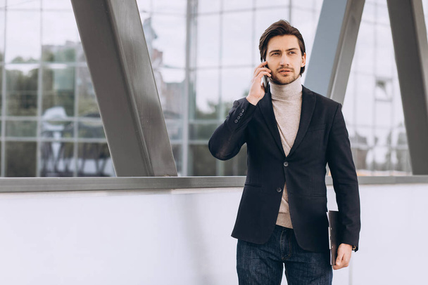 Portret przystojnego nowoczesnego mężczyzny biznesmena w garniturze trzymającego teczkę z dokumentami i rozmawiającego przez telefon na tle miejskich budynków i biur. - Zdjęcie, obraz