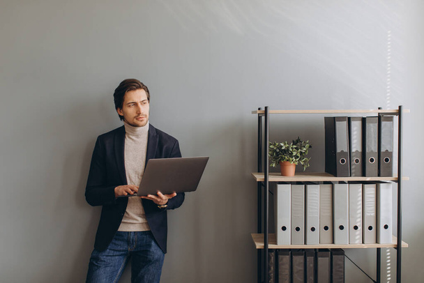 Obrázek mladého veselého podnikatele v obleku držení a používání notebooku v moderní kanceláři - Fotografie, Obrázek