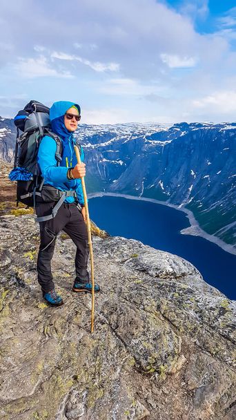 Un giovane uomo con una giacca blu e un enorme zaino da trekking si erge su una roccia, tenendo in mano un bastone. Ammira la vista di fronte a lui alte montagne innevate e un lago blu navy. - Foto, immagini