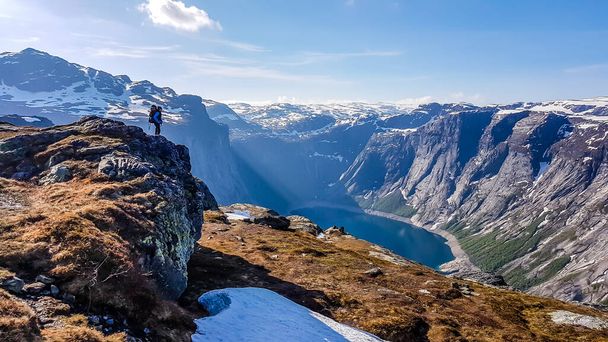青いジャケットと巨大なハイキングバックパックを着た若い男が背の高い岩の上に立って、彼の前の景色を賞賛します-背の高い雪をかぶった山々とネイビーブルーの湖。サンビームが谷に来る. - 写真・画像