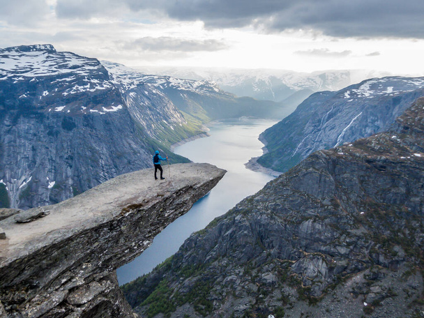 A man wearing blue jacket stands at the hanging rock formation, Trolltunga with a view on Ringedalsvatnet lake, Norway. Slopes of the mountains are partially covered with snow. Freedom and happiness - Foto, afbeelding