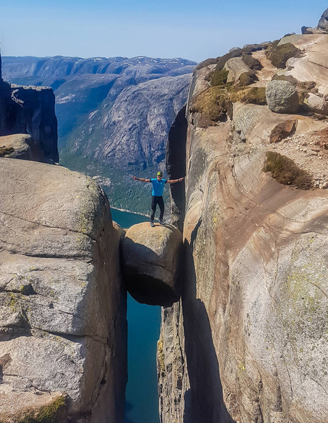 A young man wearing blue T-shirt standing on famous rock, Kjerag, Norway. The ball-shaped rock is hanging in between two stone walls with a free fall of 1000m. Boy spreads arms in a gesture of triumph - Zdjęcie, obraz