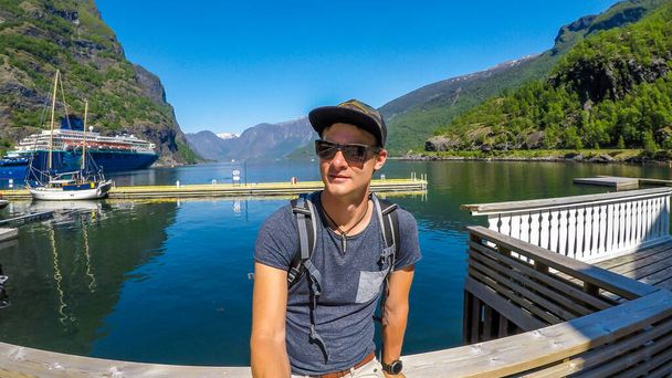 Un joven con gorra completa se toma una selfie con la pintoresca vista sobre el puerto situado en el Sognefjord, Noruega. Pocos barcos estacionados en el puerto. Reflejos de las montañas en el agua. Claro, cielo azul. - Foto, imagen