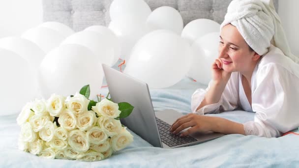 Woman in white clothes and towel on her head lying on a bed with white color balloons and bouquet of roses. Woman works at home, using her laptop, computer. Birthday, wedding. Copy space. Preparation. - Video, Çekim
