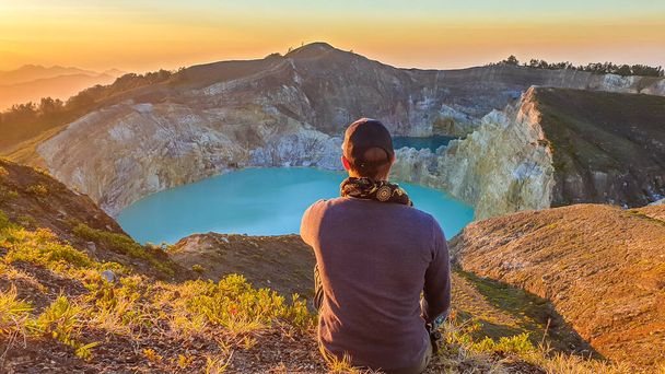 A man watching the sunrise over the Kelimutu volcanic crater lakes in Moni, Flores, Indonesia. Skyline is orange, sun slowly rising. Man is relaxed and calm, enjoying the view on turquoise lake - Foto, Imagen