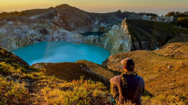 A man watching the sunrise over the Kelimutu volcanic crater lakes in Moni, Flores, Indonesia. Skyline is orange, sun slowly rising. Man is relaxed and calm, enjoying the view on turquoise lake - Foto, imagen