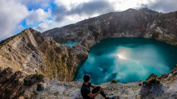 Man sitting at the volcano rim and watching the Kelimutu volcanic crater lakes in Moni, Flores, Indonesia. Man is relaxed and calm, enjoying the view on lake shining with many shades of turquoise - Foto, imagen
