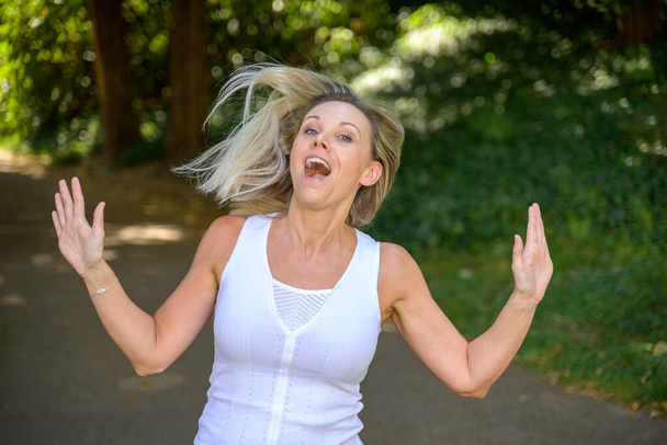 Joyful blond woman tossing her long hair and cheering outdoors in a lush green park - Foto, afbeelding