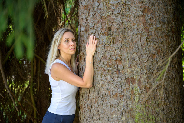 Mooie blonde vrouw staan knuffelen een boom in een park met een serieuze uitdrukking in een conservering concept - Foto, afbeelding