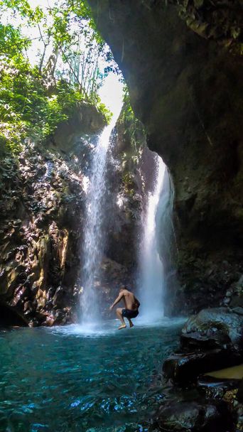 Egy férfi fürdőruhában leugrik a csúszós sziklákról a Gitgit Twin Waterfall előtt, Balin, Indonéziában. A vízesést sziklák veszik körül. A férfi, aki élvezi a természet szépségét. Kaland - Fotó, kép