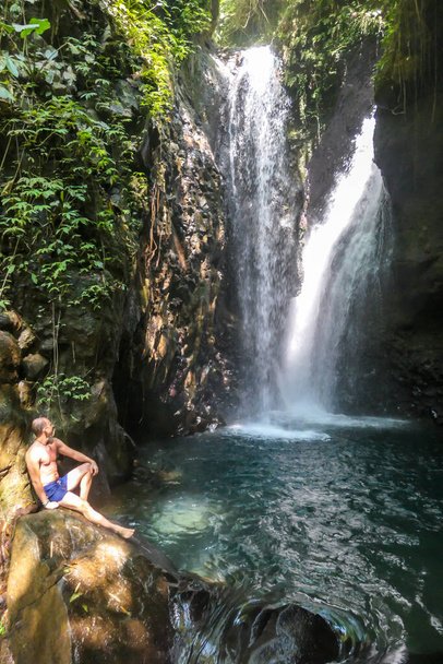Un uomo in costume da bagno seduto sulle rocce di fronte a Gitgit Twin Waterfall, Bali, Indonesia. La cascata è circondata da rocce. L'uomo che gode della bellezza della natura. Avventura e scoperta - Foto, immagini