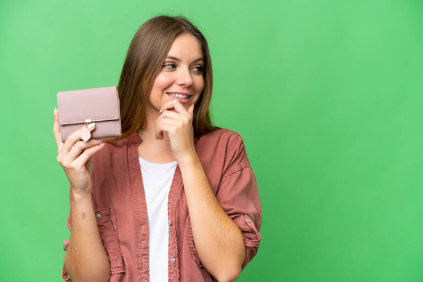 Νεαρή ξανθιά γυναίκα κρατώντας ένα πορτοφόλι πάνω από απομονωμένο φόντο chroma κλειδί σκέφτεται μια ιδέα και ψάχνει πλευρά - Φωτογραφία, εικόνα