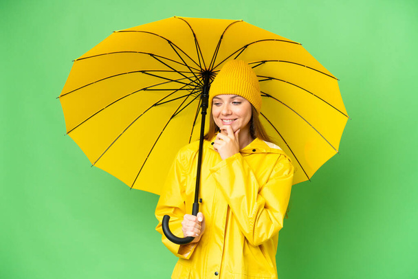 Молодая блондинка с непромокаемым плащом и зонтиком на изолированном фоне хрома ключ, глядя в сторону и улыбаясь - Фото, изображение